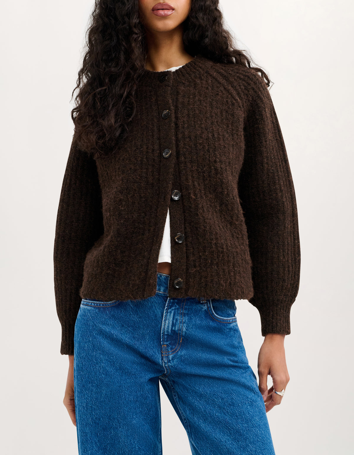 A woman wearing a dark brown textured cable-knit cardigan with a button-up front, paired with blue jeans, against a plain white studio background.