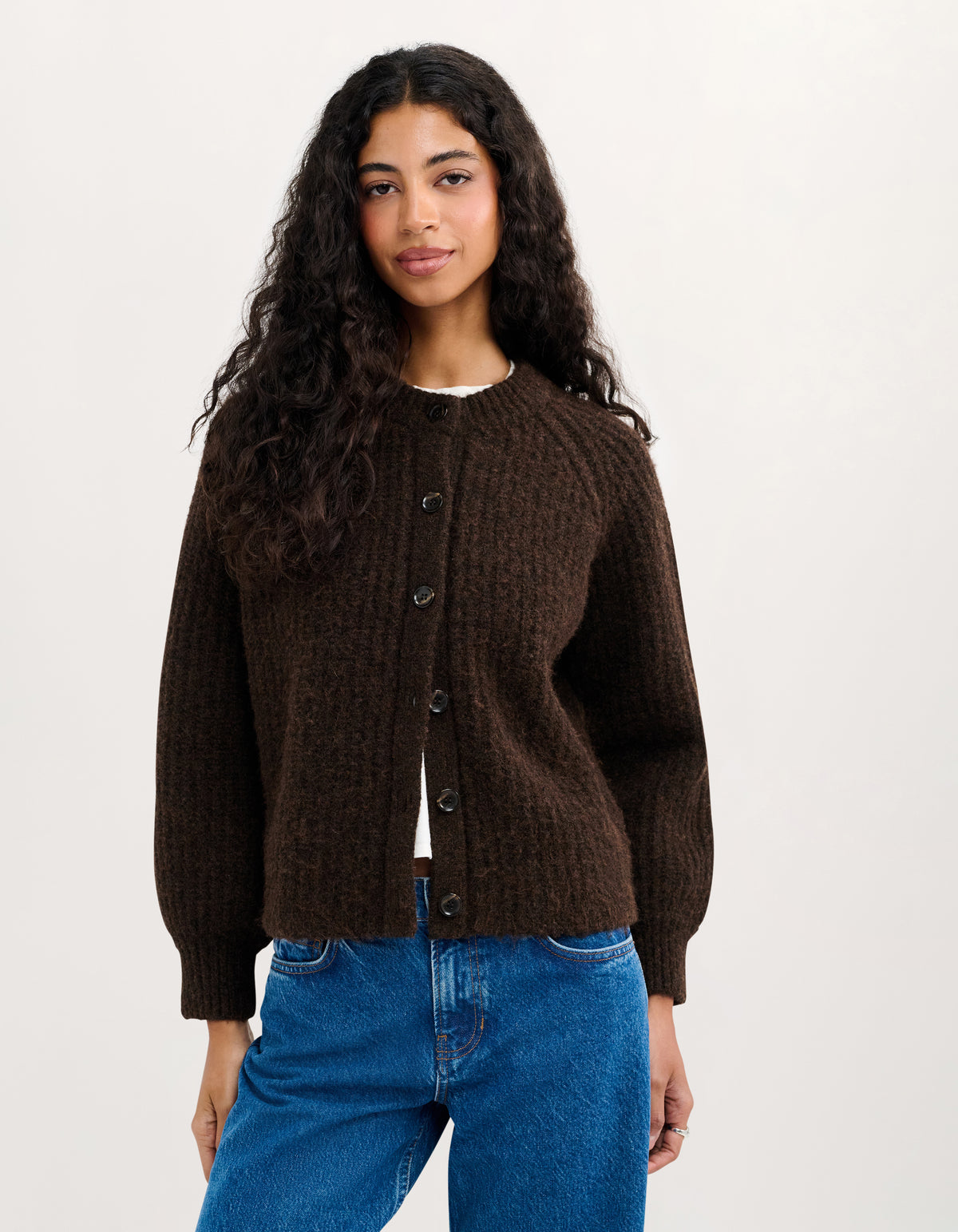 A woman with long, curly dark hair wearing a dark brown textured cable-knit cardigan sweater and blue jeans, standing against a plain off-white studio background.