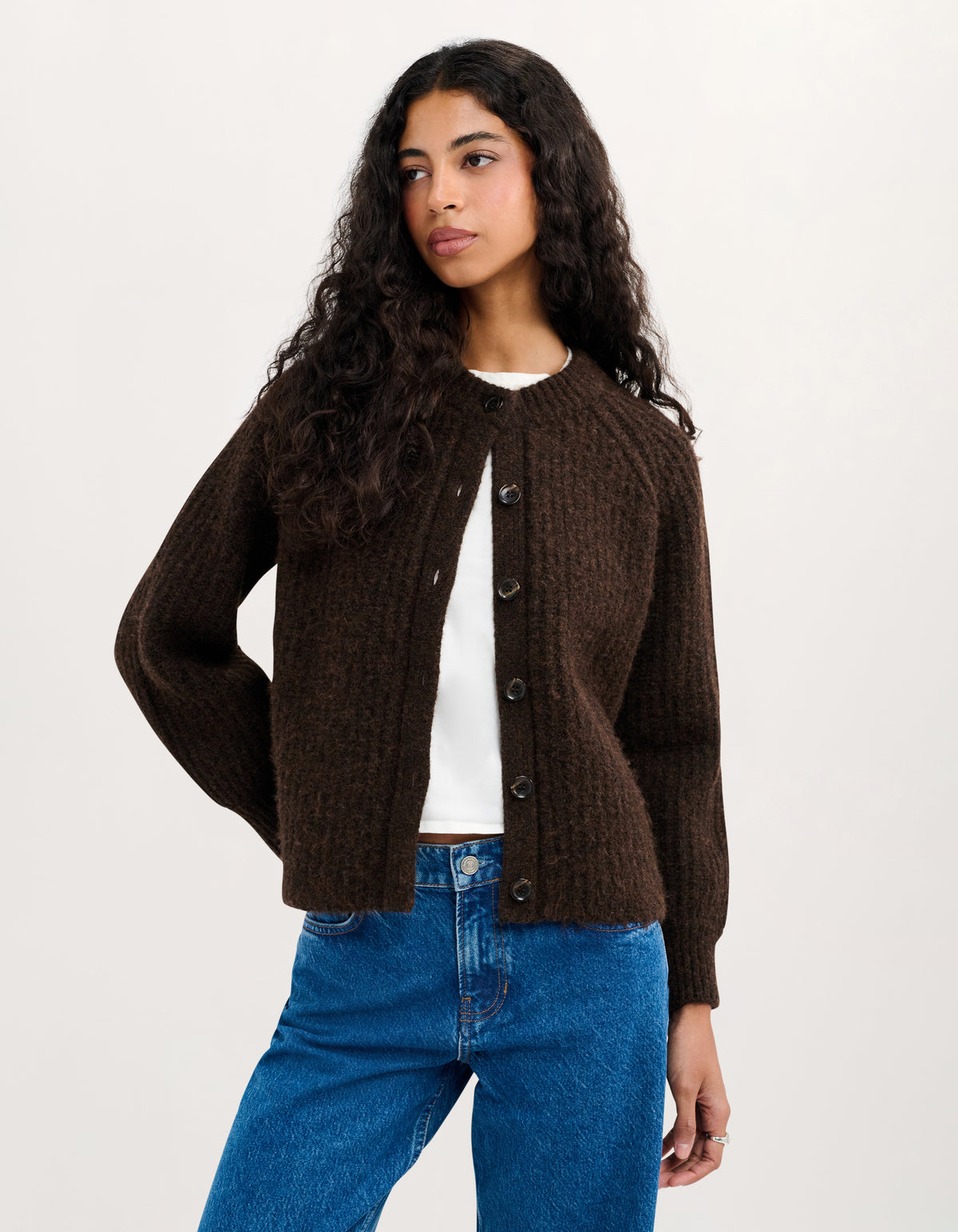 A woman modeling a dark brown textured knit cardigan with dark buttons, worn open over a white t-shirt and blue jeans, against a plain light background.