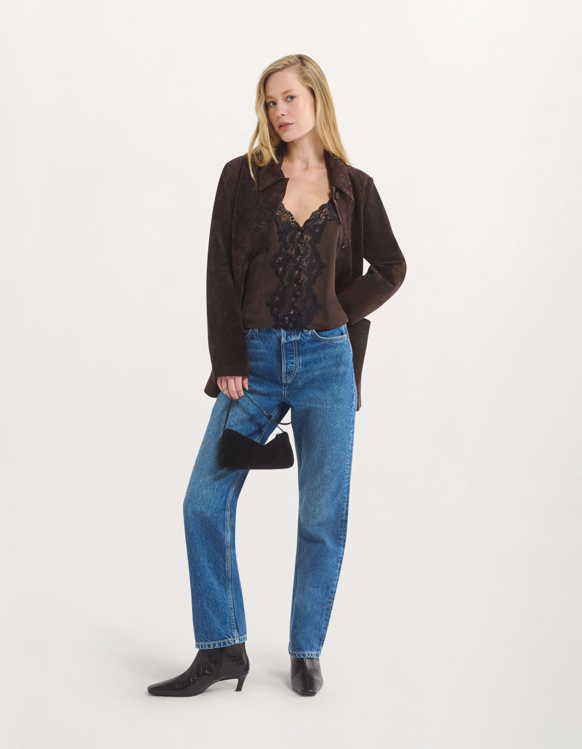 A blonde model stands against a light gray studio background wearing a dark brown suede jacket over a black lace top, blue jeans, and black ankle boots, while holding a small black handbag.