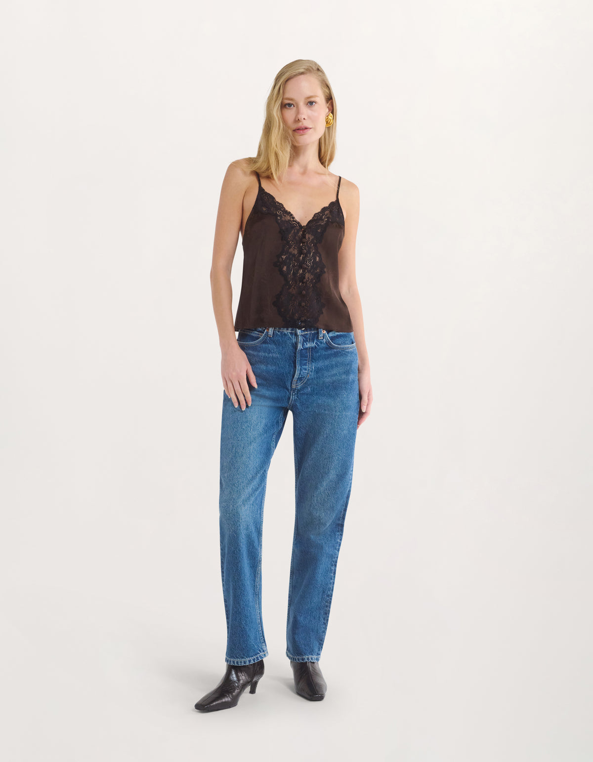 A blonde model stands against a plain light gray studio background wearing a dark brown camisole top with black lace detailing, blue jeans, and black ankle boots.