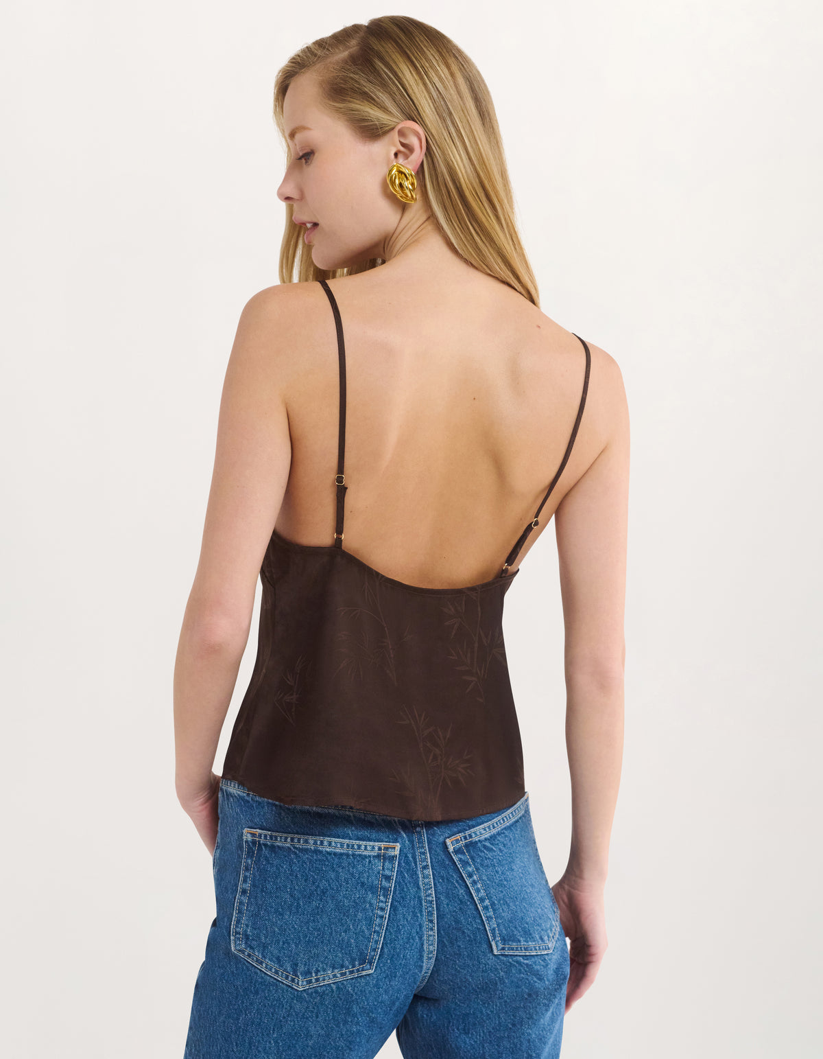 A woman shown from the back wearing a dark brown spaghetti-strap camisole top with a subtle tonal damask pattern, paired with blue jeans, against a plain off-white background.