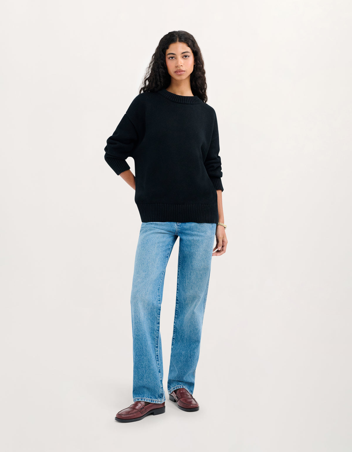 A woman modeling a black crewneck sweater, light-wash blue jeans, and dark brown leather loafers against a plain off-white studio background.