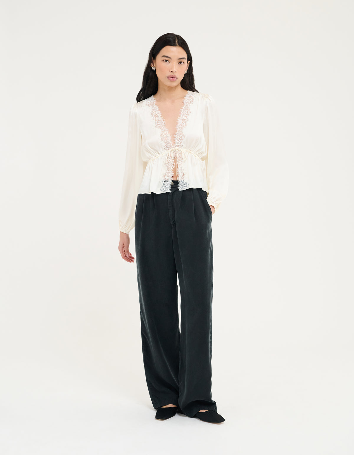 A model stands against a plain white studio background wearing a white long-sleeve lace-trim blouse with a tie waist and black wide-leg trousers.