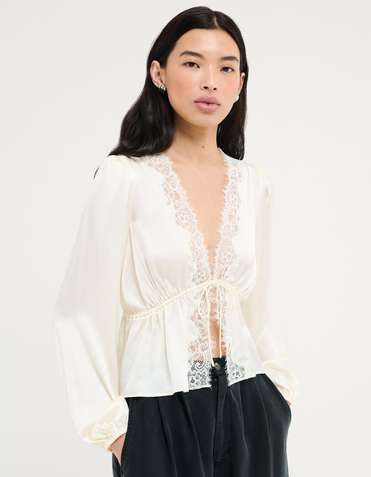 A woman modeling a cream satin long-sleeve blouse with a lace V-neckline and drawstring waist, paired with black pants, against a plain off-white background.