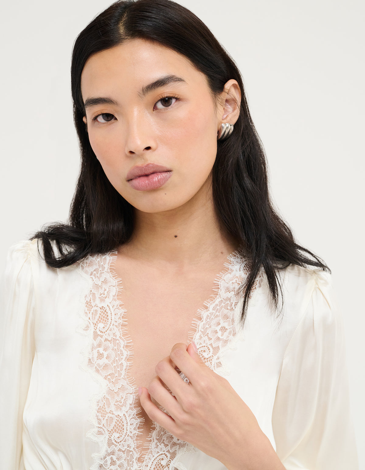 A woman with long dark hair wearing a white satin blouse with a lace V-neckline, photographed against a plain light background.
