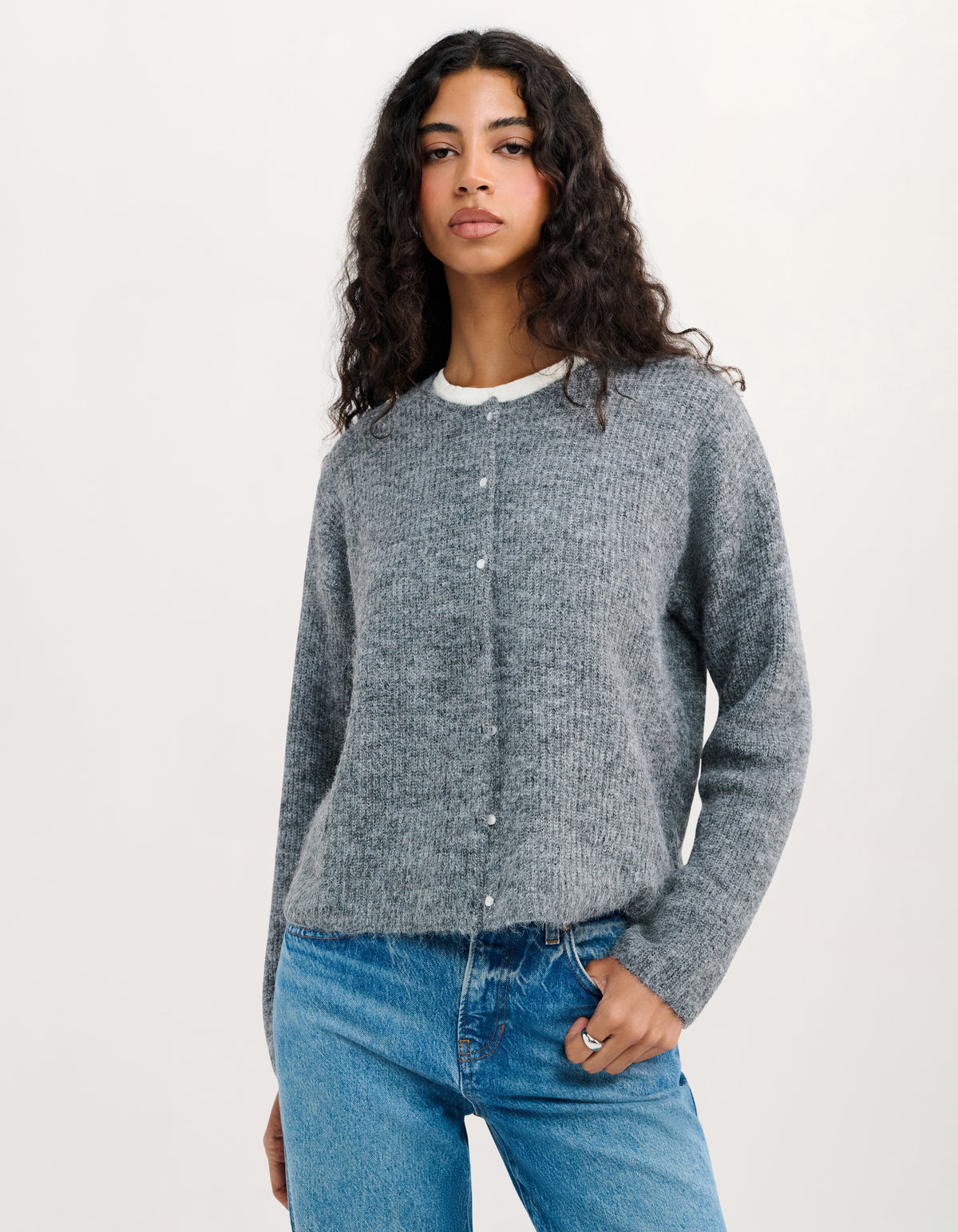 A woman with long curly dark hair wearing a heather gray button-up cardigan over a white t-shirt and blue jeans, standing against a plain off-white studio background.