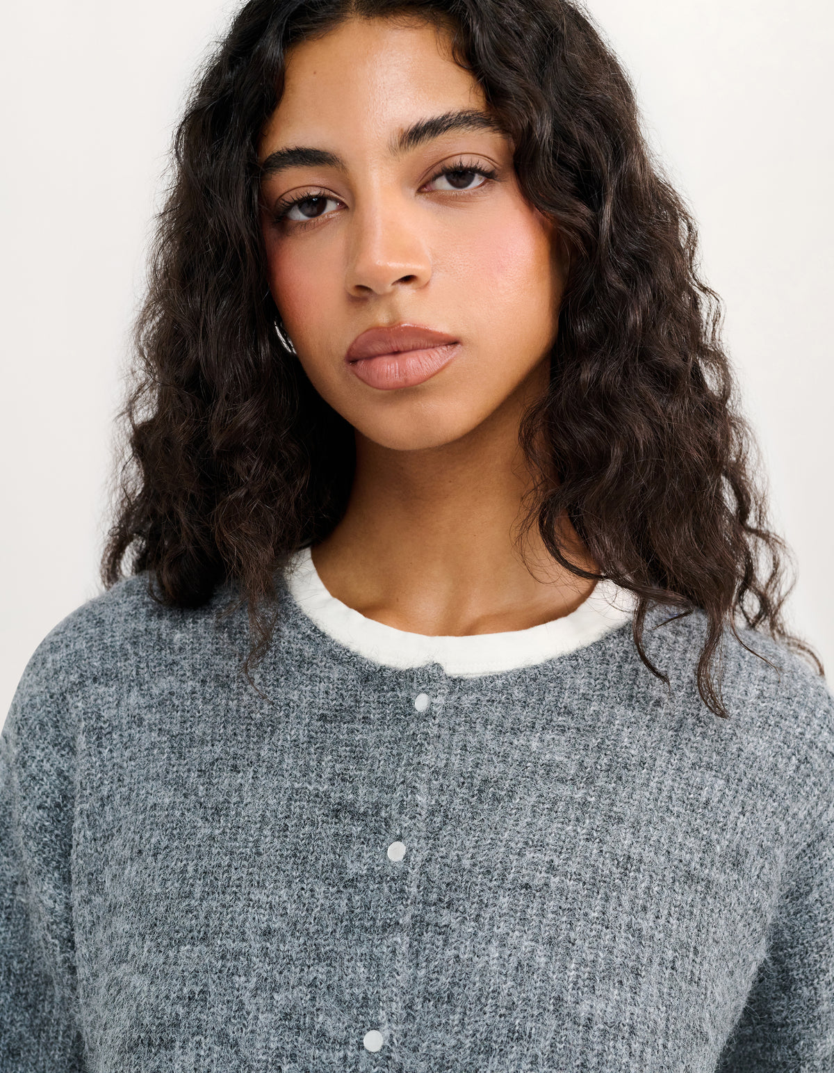 A close-up shot of a woman with curly dark hair wearing a gray textured knit cardigan with a white ribbed crew neck and white buttons, set against a plain off-white background.