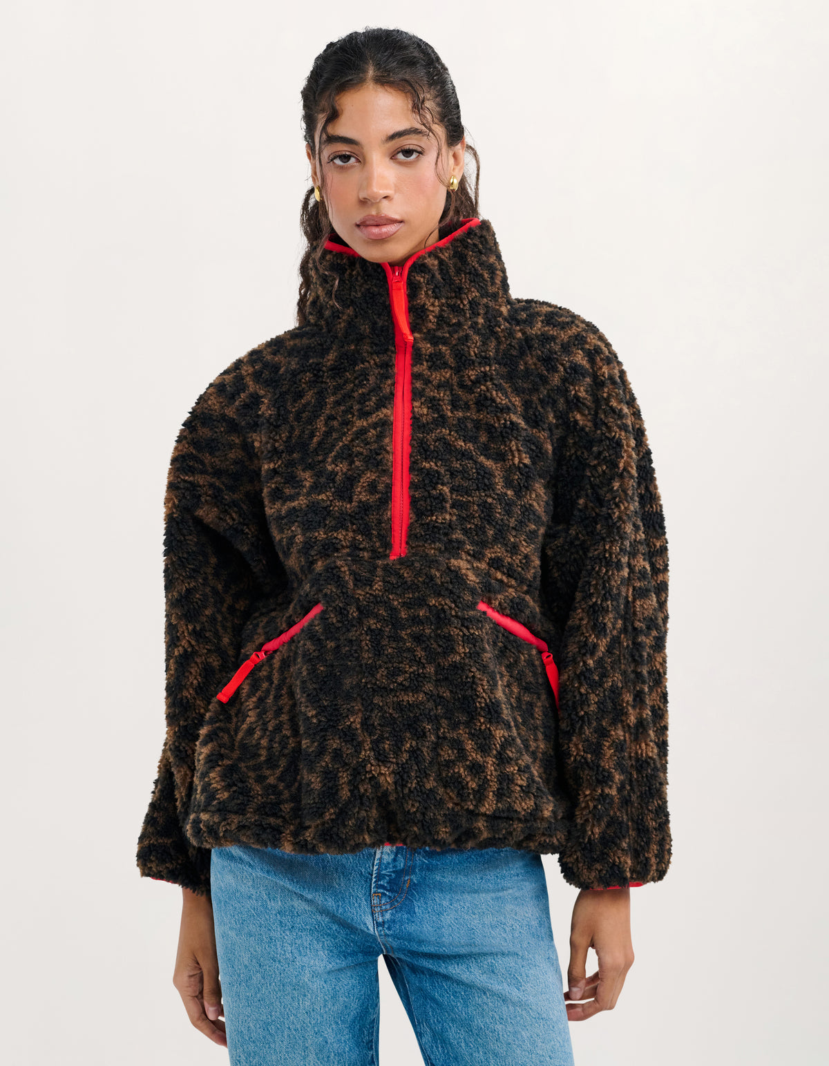 A model wearing a black and brown leopard-print fleece pullover with bright red zipper accents, paired with blue jeans, against a plain white studio background.