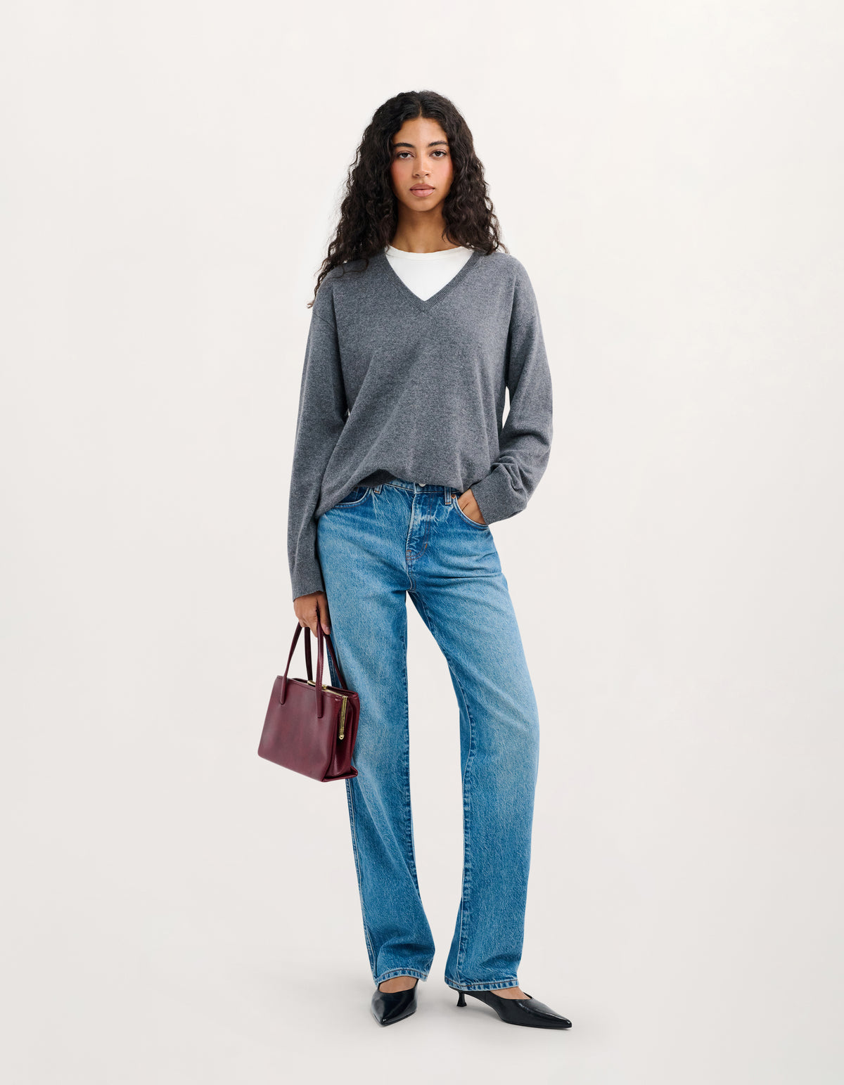 A woman stands against a plain light background wearing a gray V-neck knit sweater over a white top, blue jeans, black pointed flats, and holding a small burgundy handbag.