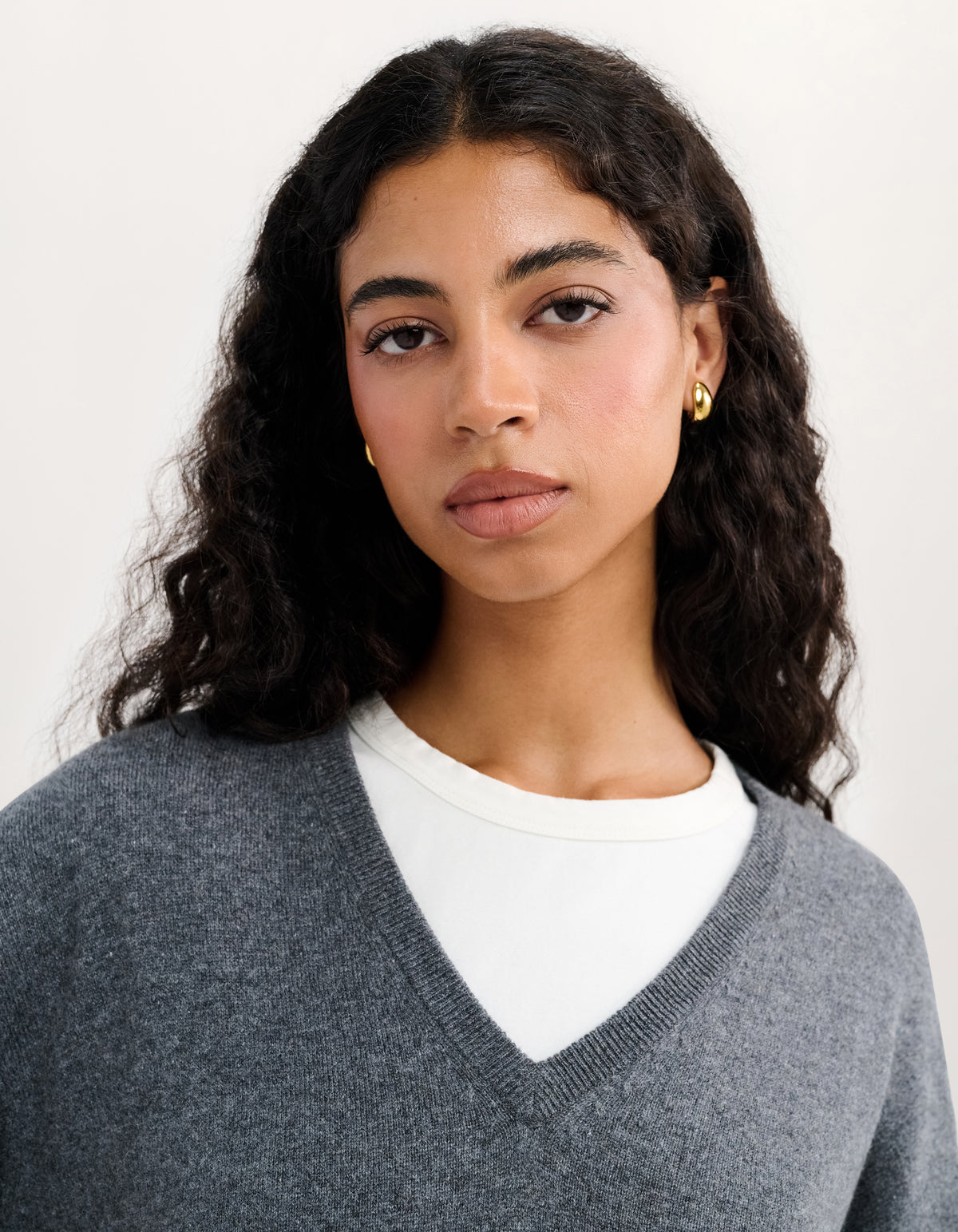 A woman with dark curly hair wearing a gray V-neck knit sweater over a white crewneck top, with small gold hoop earrings, photographed against a plain light background.