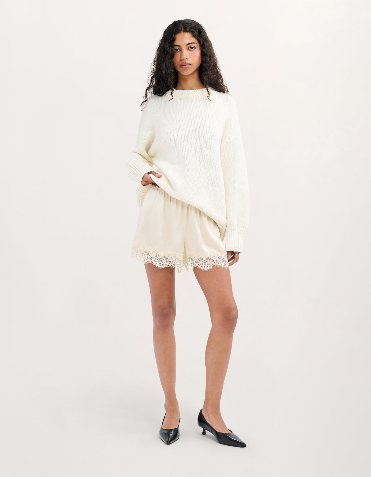 A model stands against a plain light background wearing a cream-colored knit sweater and matching shorts with lace trim, paired with black pointed-toe heels.