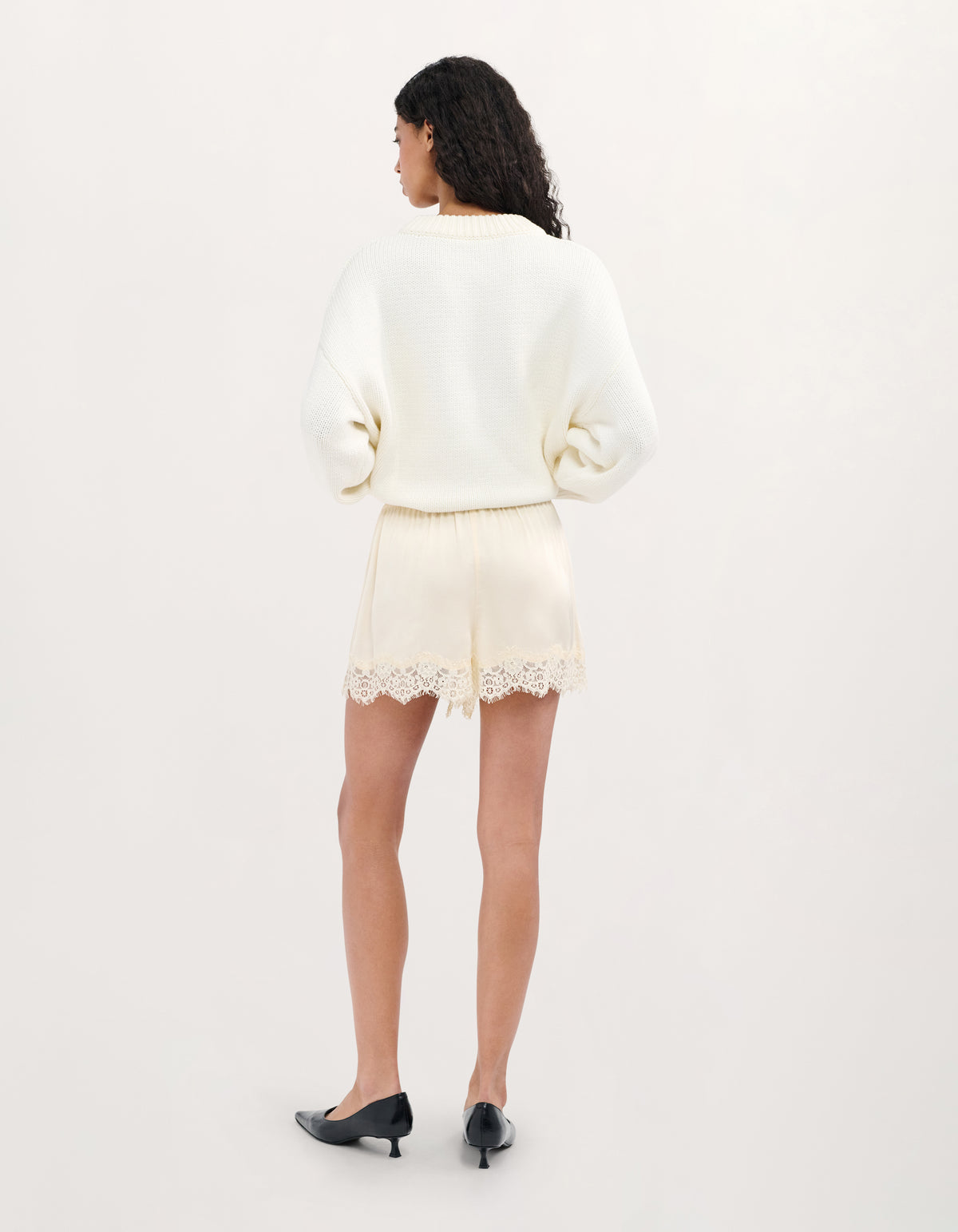 A model is shown from the back wearing a cream-colored knit sweater and matching cream lace-trimmed shorts, paired with black pointed-toe heels, against a plain off-white studio background.