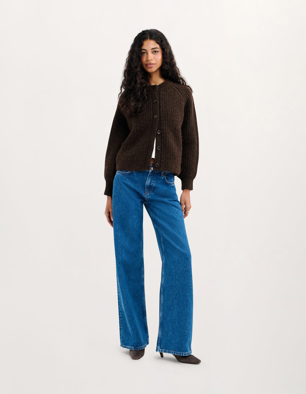 A woman modeling a dark brown button-up knit cardigan and wide-leg blue jeans against a plain light gray studio background.