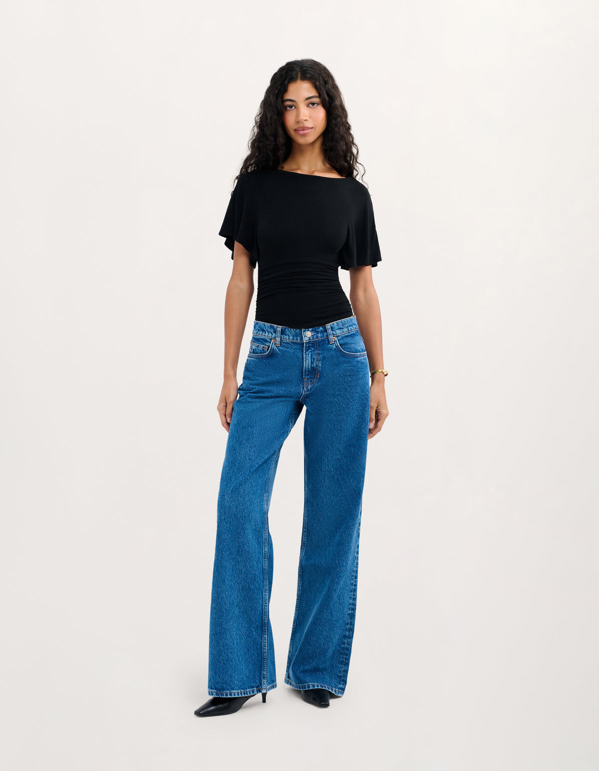 A woman modeling a black short-sleeve top and blue wide-leg jeans against a plain light gray studio background.