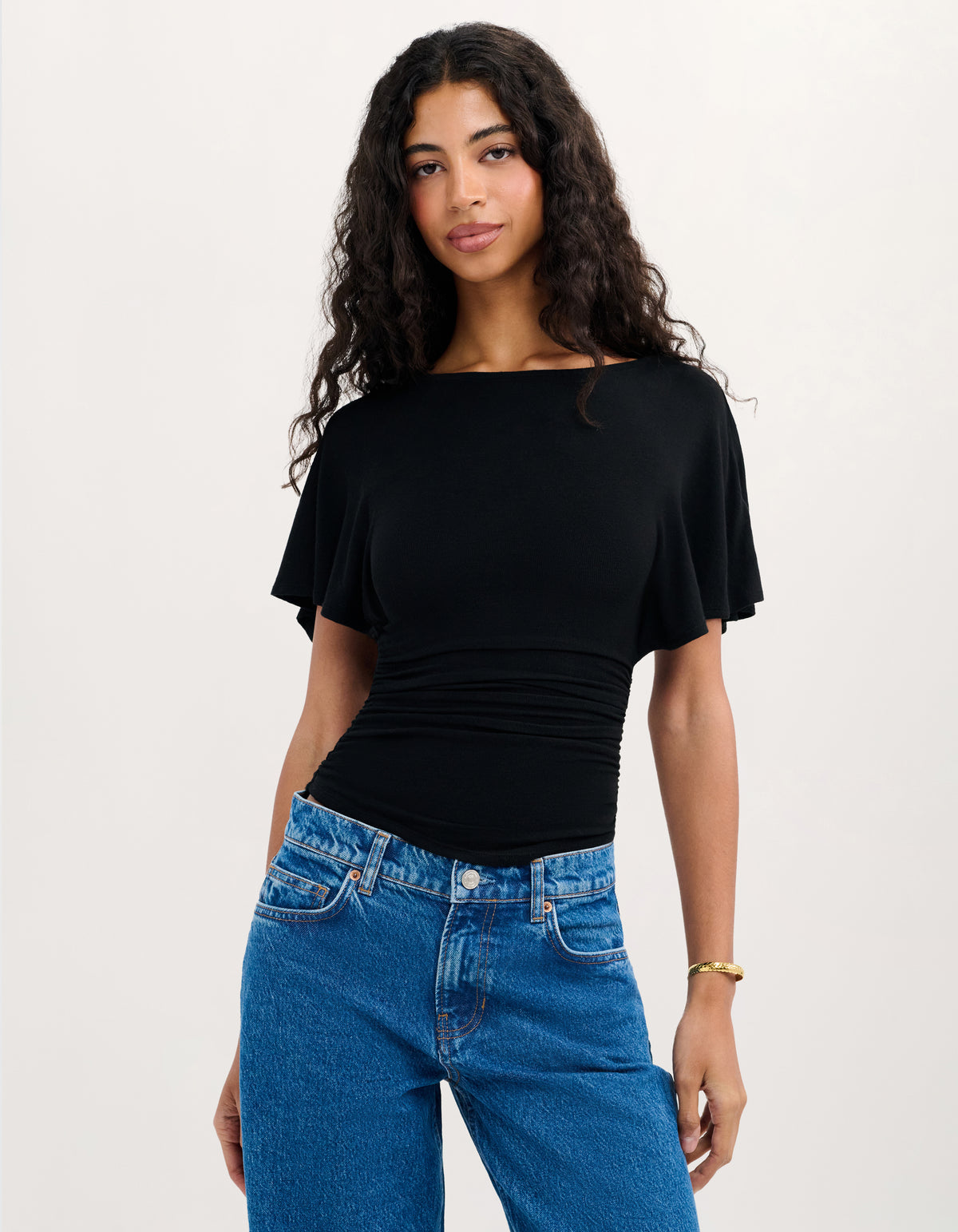 A woman modeling a black short-sleeve top with a boat neckline and flared sleeves, paired with blue jeans, against a plain light background.
