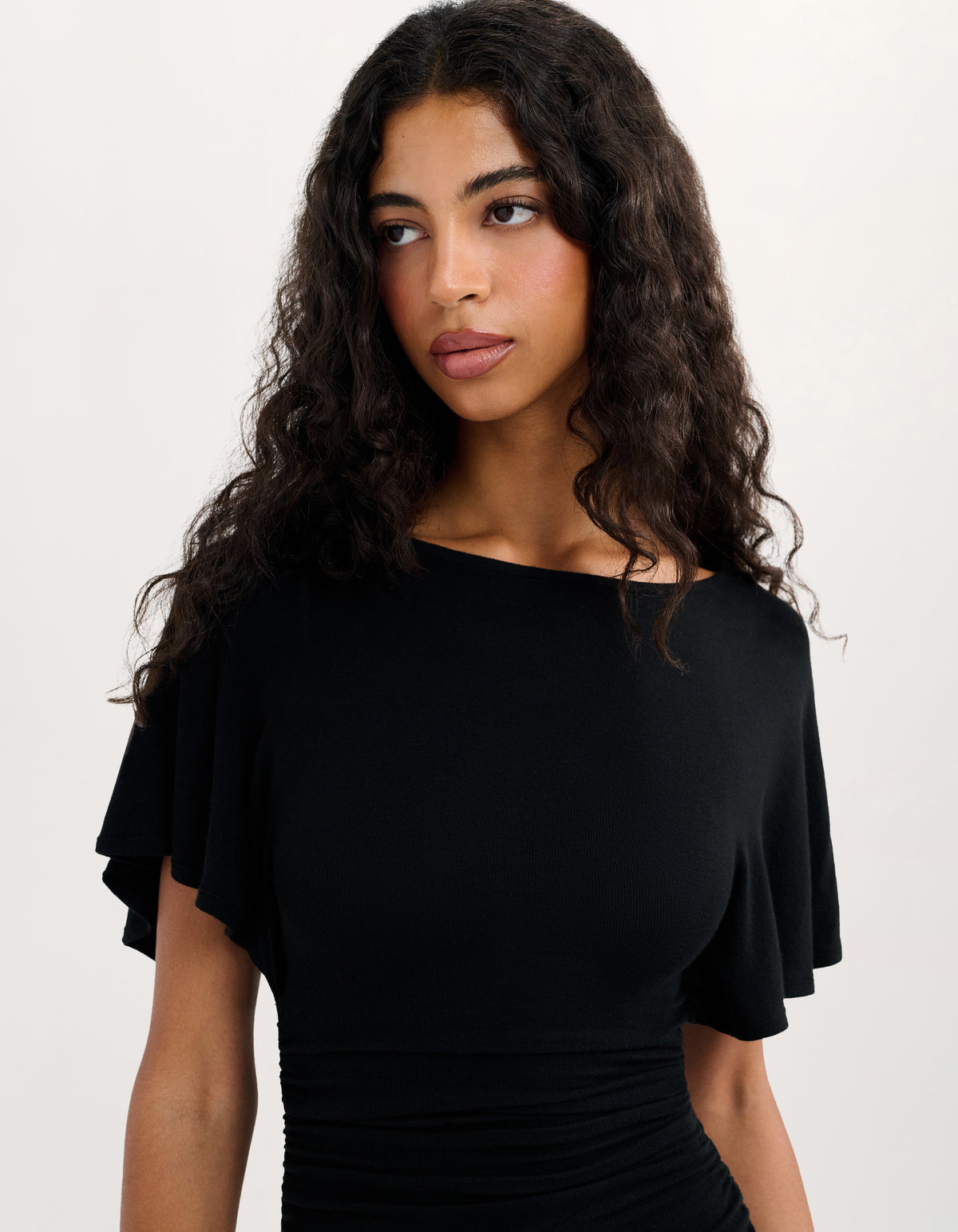 A woman with long, curly dark hair wearing a black off-the-shoulder top with fluttery ruffle sleeves and a ruched waist, photographed against a plain light background.