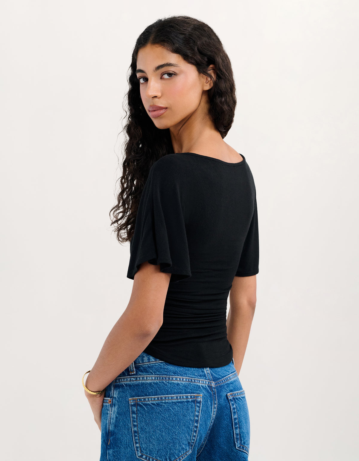 A woman wearing a black short-sleeve top with flutter sleeves and blue jeans, photographed from the back against a plain light background.