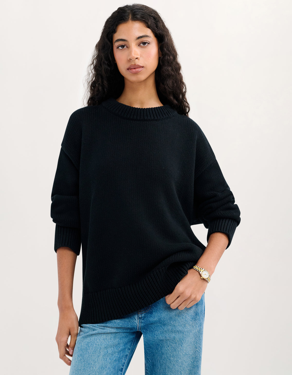 A woman modeling a black knit crewneck sweater with three-quarter sleeves, paired with blue jeans, against a plain off-white studio background.