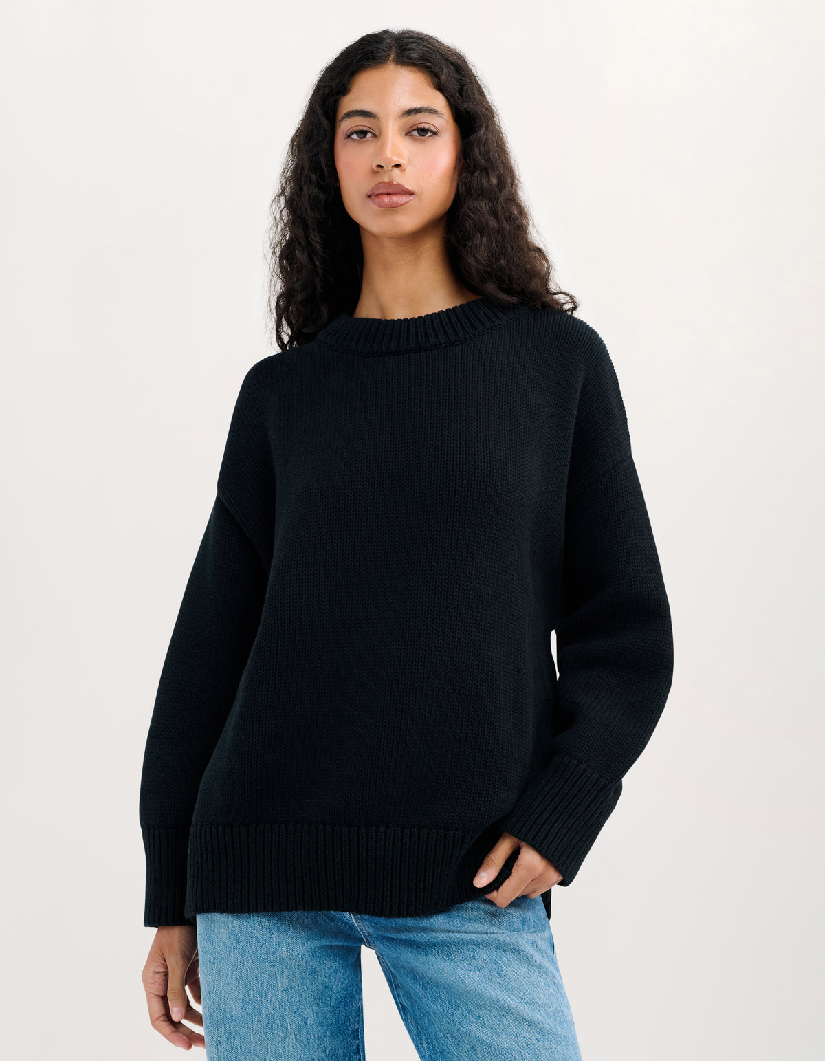 A woman wearing a black ribbed knit crewneck sweater and light blue jeans, standing against a plain white studio background.