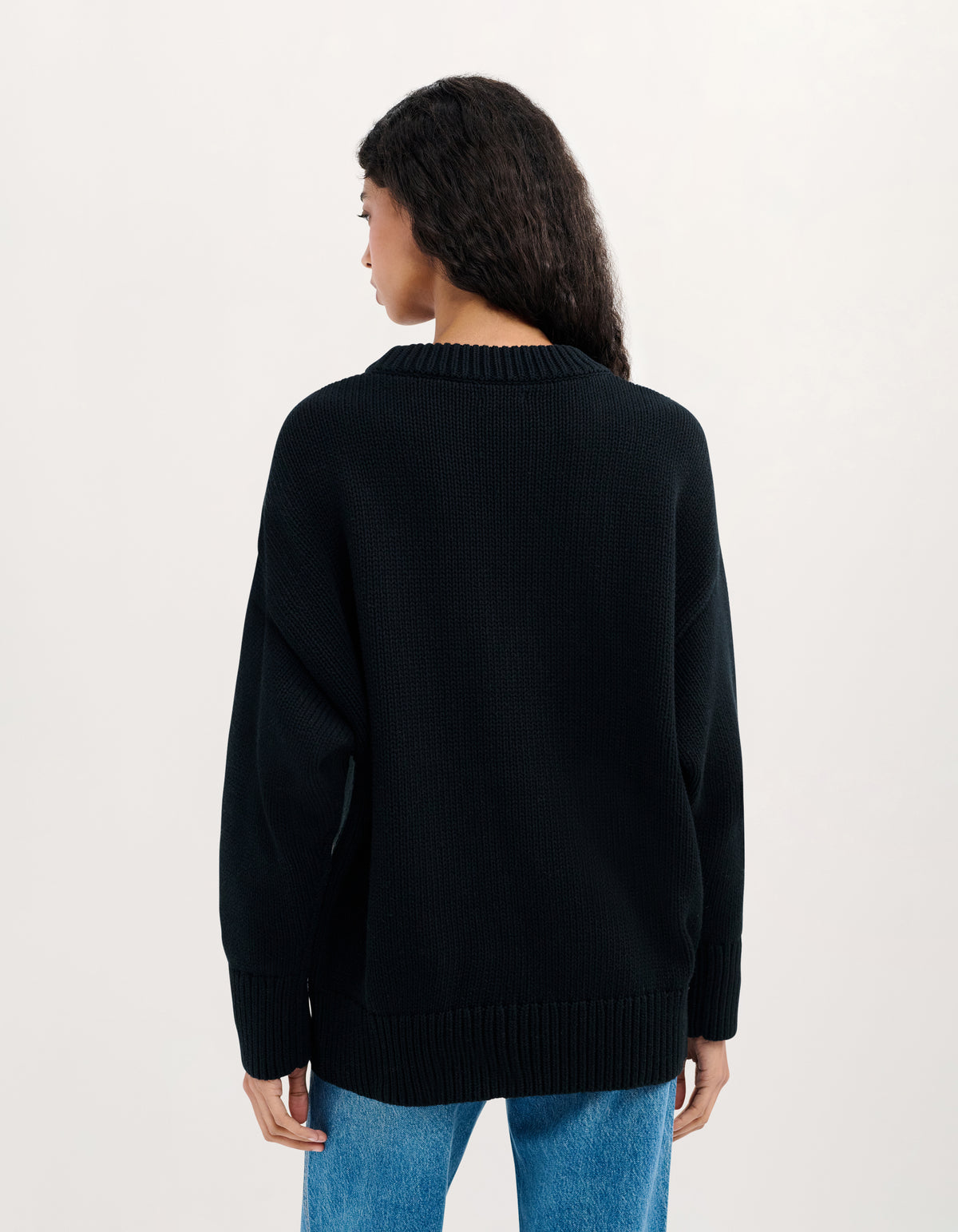 A woman viewed from the back wearing a black ribbed knit sweater and blue jeans against a plain light background.