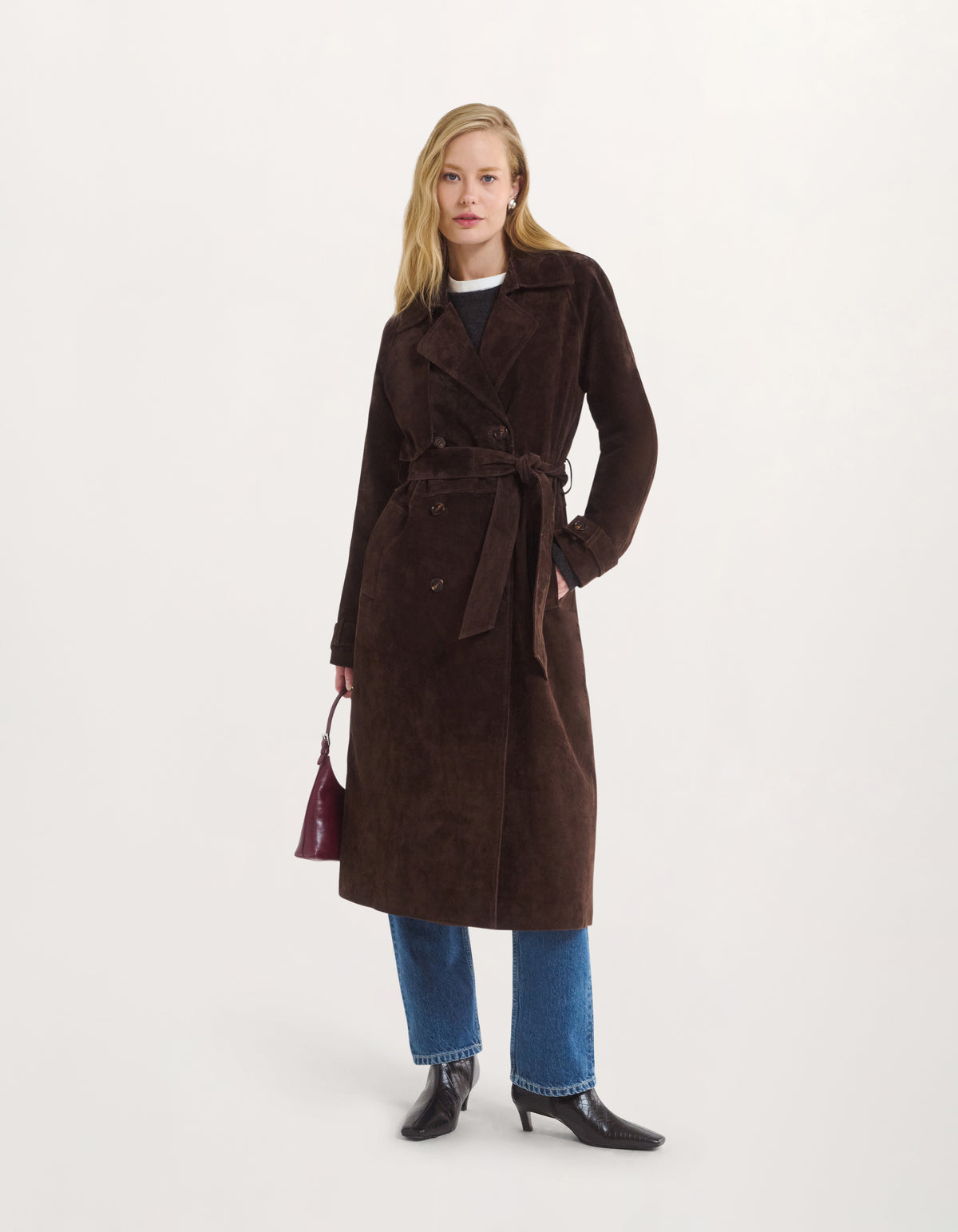 A woman models a long, dark brown suede belted coat over a black top and blue jeans, holding a small burgundy handbag against a plain off-white studio background.