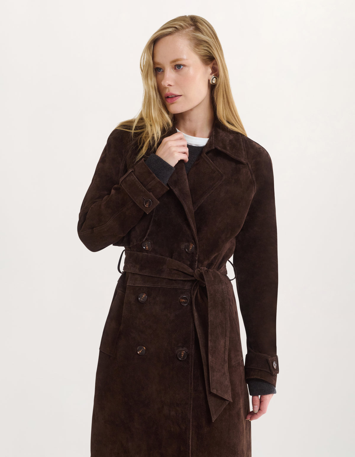 A woman modeling a dark brown suede double-breasted trench coat with a matching waist tie, worn over a black top, against a plain white studio background.