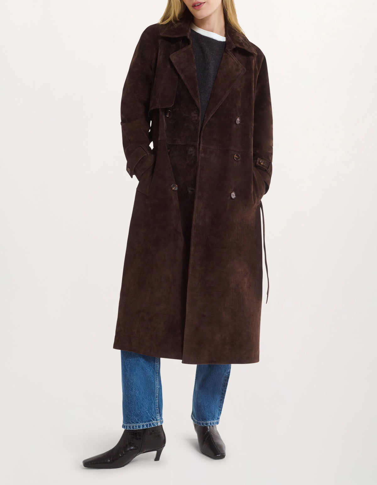 A model wearing a long, double-breasted brown suede trench coat over a dark top, blue jeans, and black ankle boots, photographed against a clean white studio background.
