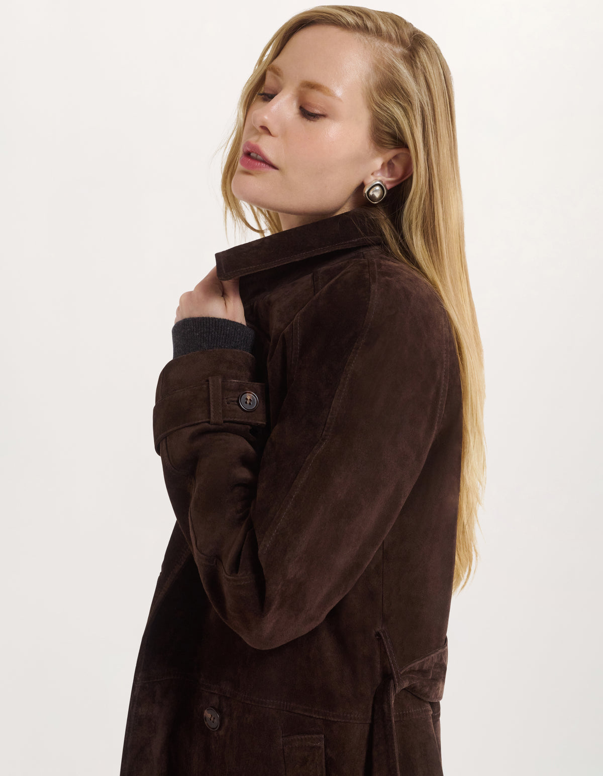 A blonde woman wearing a dark brown suede jacket with a high collar, shown in a side profile against a plain white studio background.