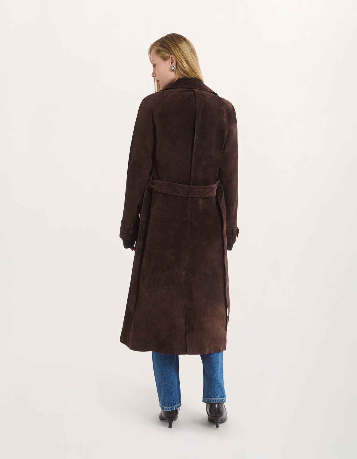 A woman viewed from the back wearing a long dark brown suede-like coat with a matching waist belt, paired with blue jeans and black shoes, against a plain light gray studio background.