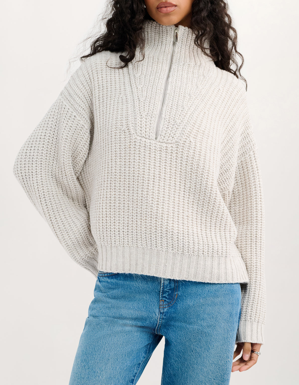 A woman wearing a chunky knit quarter-zip sweater in a light oatmeal beige color, paired with blue jeans, against a plain white studio background.
