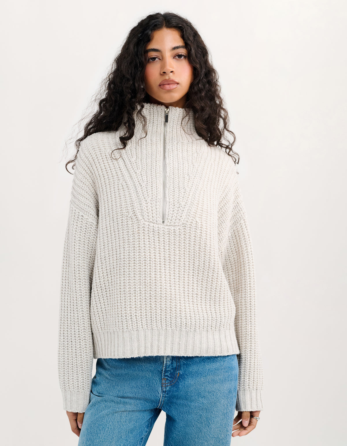 A woman with long, curly dark hair wearing a cream-colored, textured half-zip mock-neck sweater and blue jeans, standing against a plain off-white studio background.