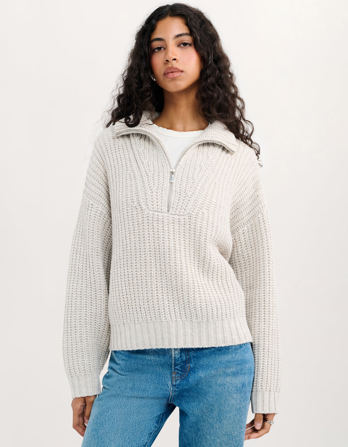 A woman with long, curly dark hair wearing a cream-colored, textured waffle-knit half-zip sweater and blue jeans, standing against a plain white studio background.