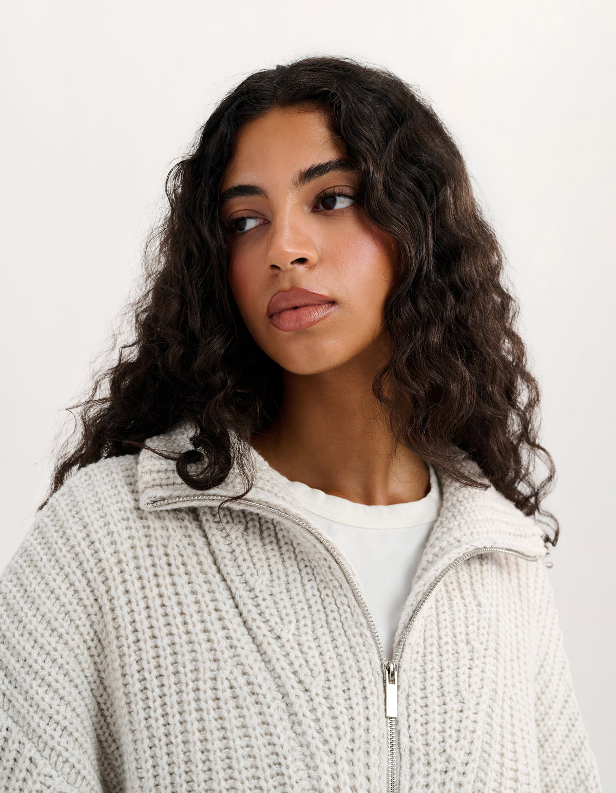 A woman with long, dark, curly hair wearing a cream-colored, textured knit zip-up hoodie over a white t-shirt, photographed against a plain off-white background.