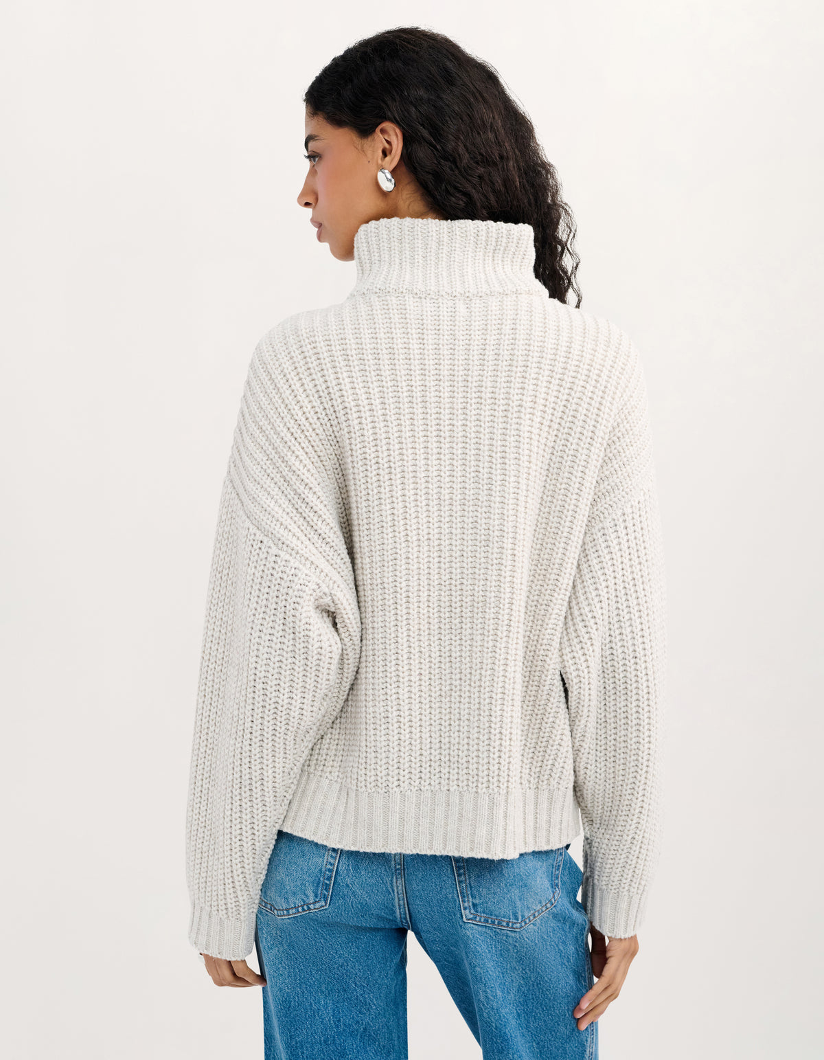 A woman shown from the back wearing a cream-colored, textured waffle-knit turtleneck sweater and blue jeans against a plain white studio background.