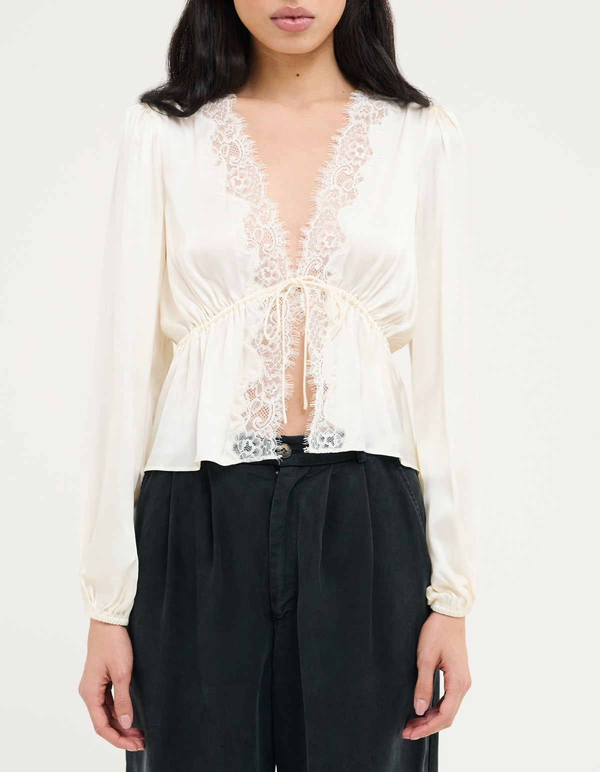 A woman wearing a cream satin long-sleeve blouse with a lace V-neckline and drawstring waist, paired with black wide-leg pants, against a plain light background.