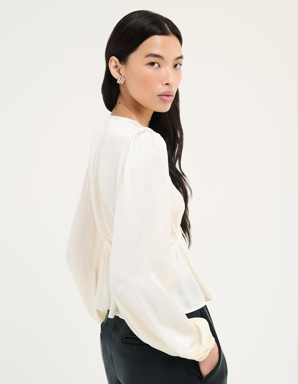 A woman wearing a cream-colored long-sleeve blouse with a gathered waist and puffed sleeves, paired with black pants, photographed against a plain white studio background.