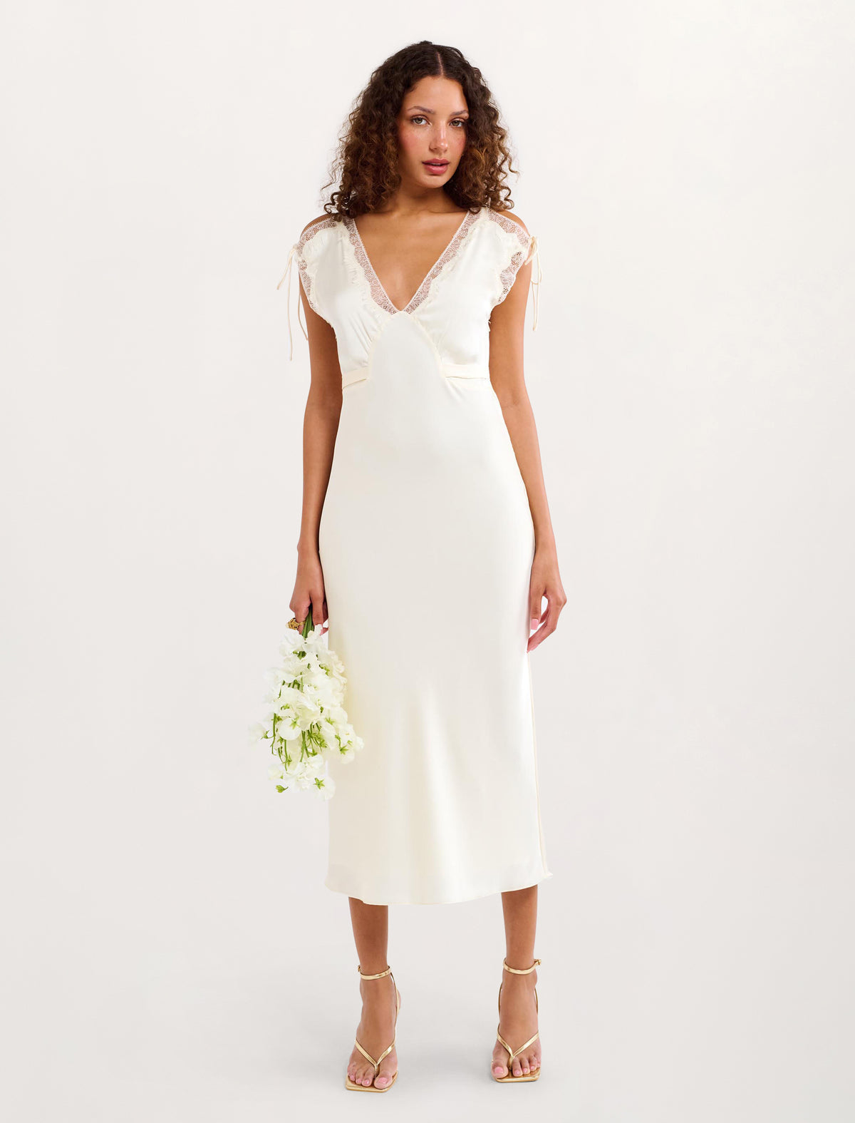 A woman modeling a white midi-length slip dress with a deep V-neckline, lace-trimmed cap sleeves, and a subtle sheen, holding a small bouquet of white flowers against a plain white studio background.