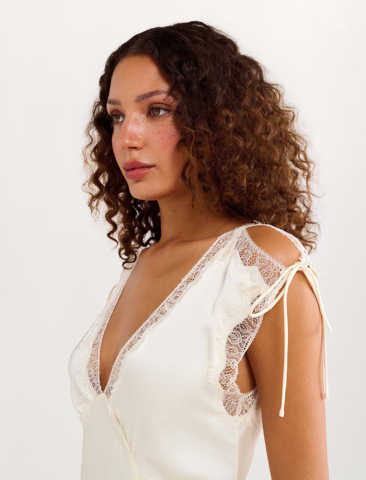 A woman with curly brown hair wearing a cream-colored lace-trimmed camisole top with tie-shoulder straps, photographed against a plain white studio background.