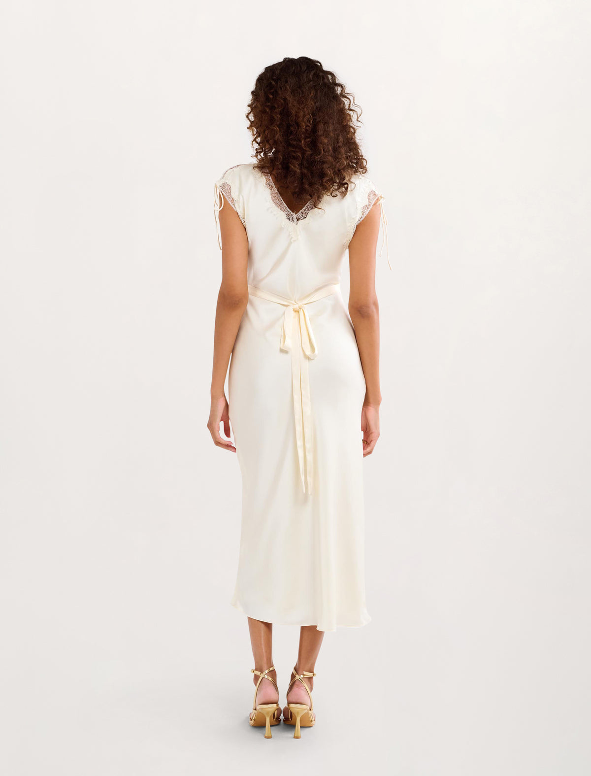 A woman viewed from the back wearing a cream-colored midi dress with a wrap-style V-neckline, short cap sleeves, and a matching fabric tie belt at the waist. She is wearing gold high-heeled sandals against a plain white studio background.