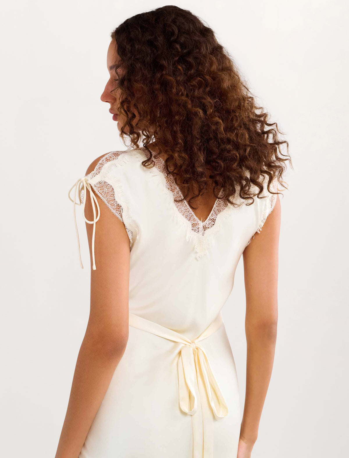 A woman with long, curly brown hair is shown from the back wearing a cream-colored dress with lace shoulder details and a matching fabric tie belt at the waist, set against a plain white studio background.