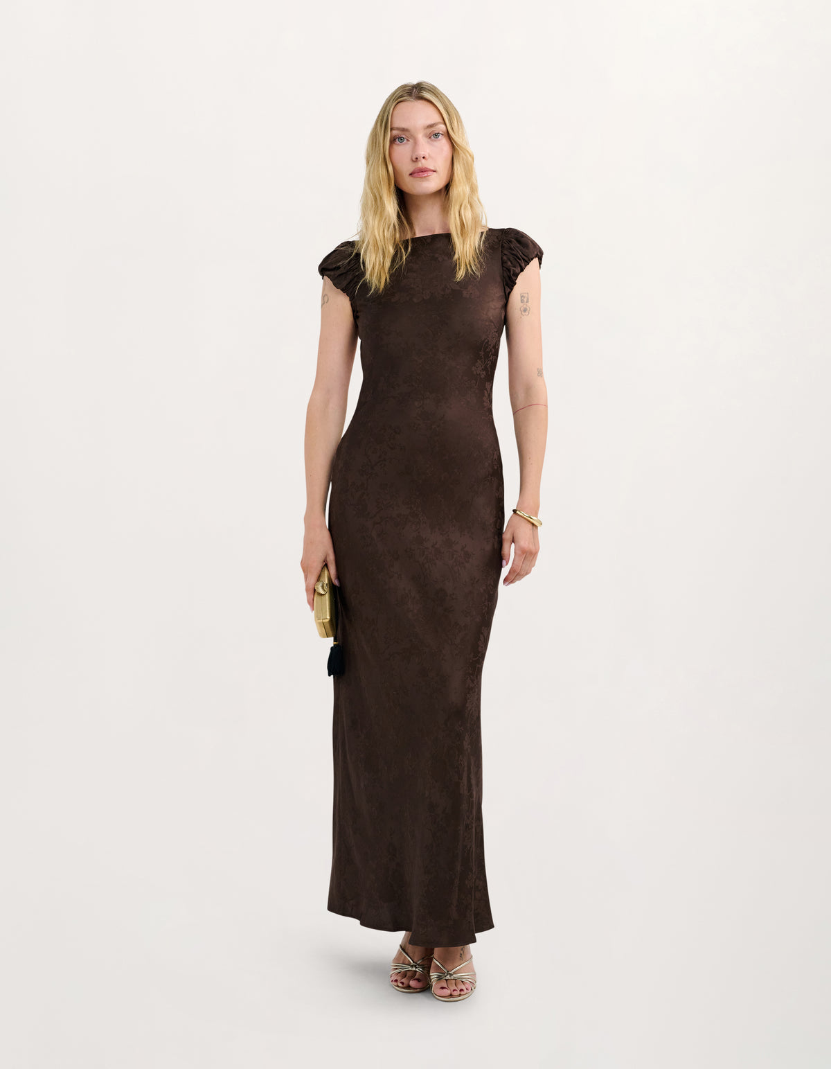 A woman modeling a dark brown, textured, sleeveless maxi dress with cap sleeves and a high neckline, standing against a plain off-white studio background.