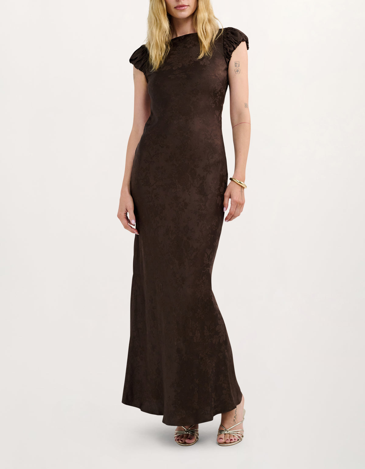 A woman modeling a dark brown, textured maxi dress with ruffled cap sleeves and a high neckline, set against a plain light gray background.
