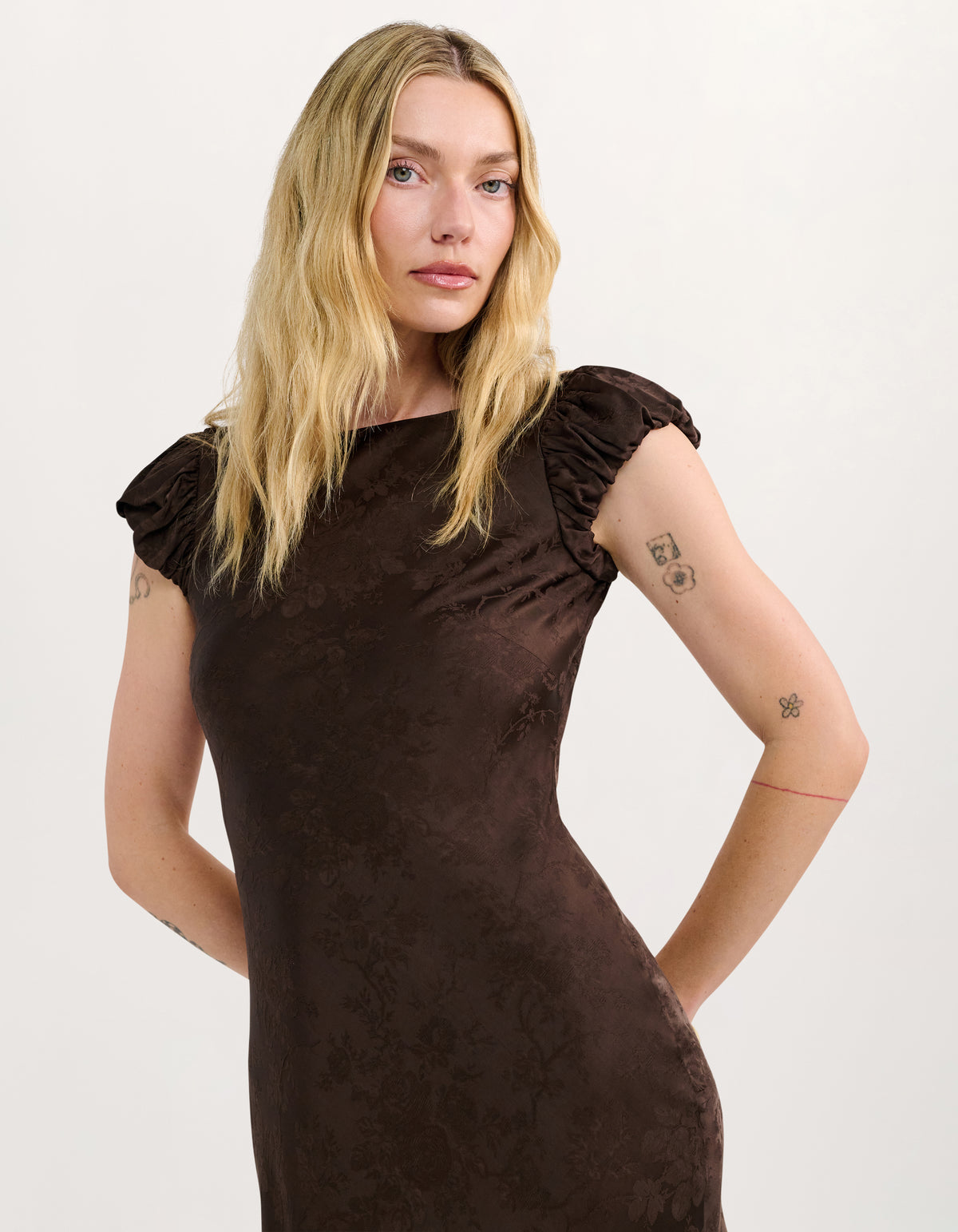 A blonde model wearing a dark brown textured dress with puffed cap sleeves, photographed against a light gray studio background.
