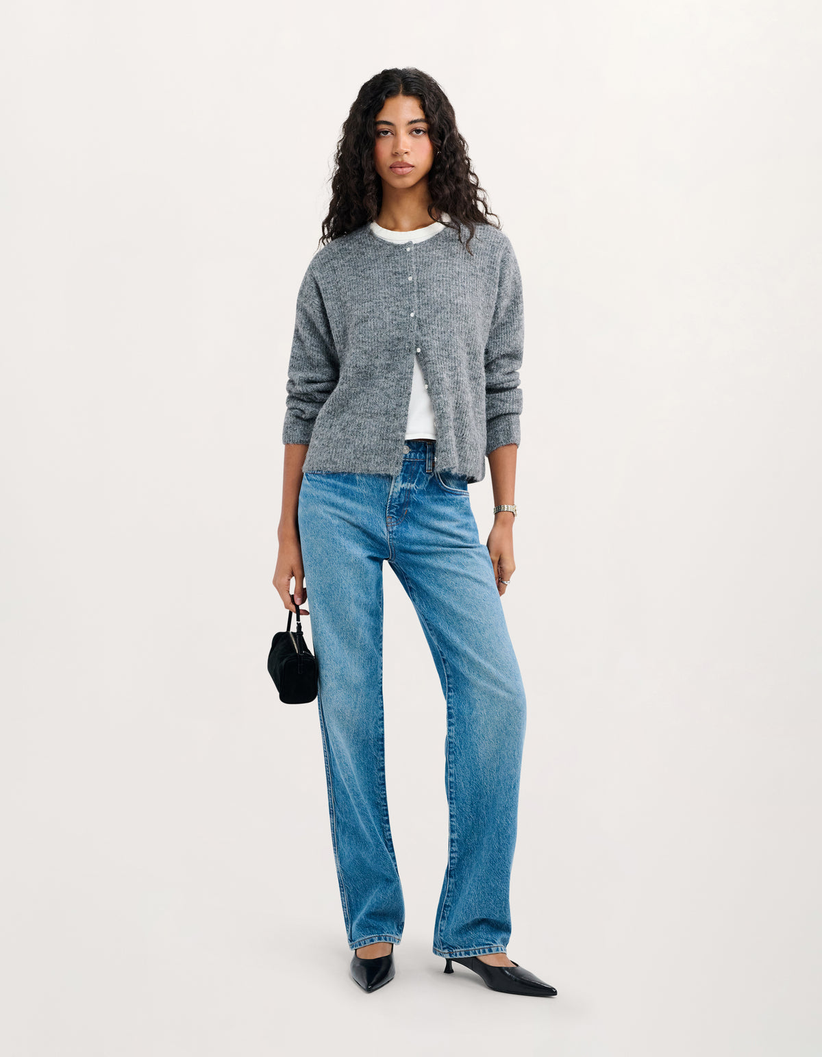 A model stands against a plain light background wearing a gray knit cardigan over a white top, blue jeans, and black pointed flats, while holding a small black handbag.