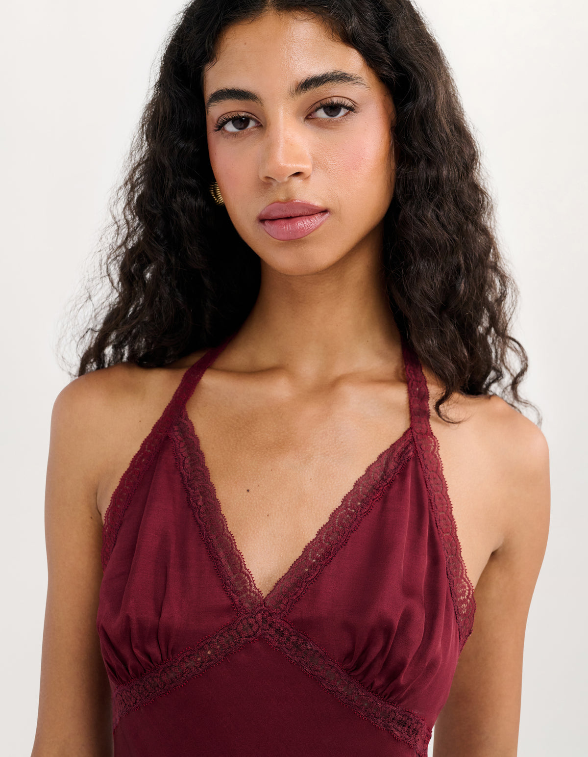 A woman with long, curly dark hair wearing a burgundy halter-neck camisole with lace trim, photographed against a clean off-white background.