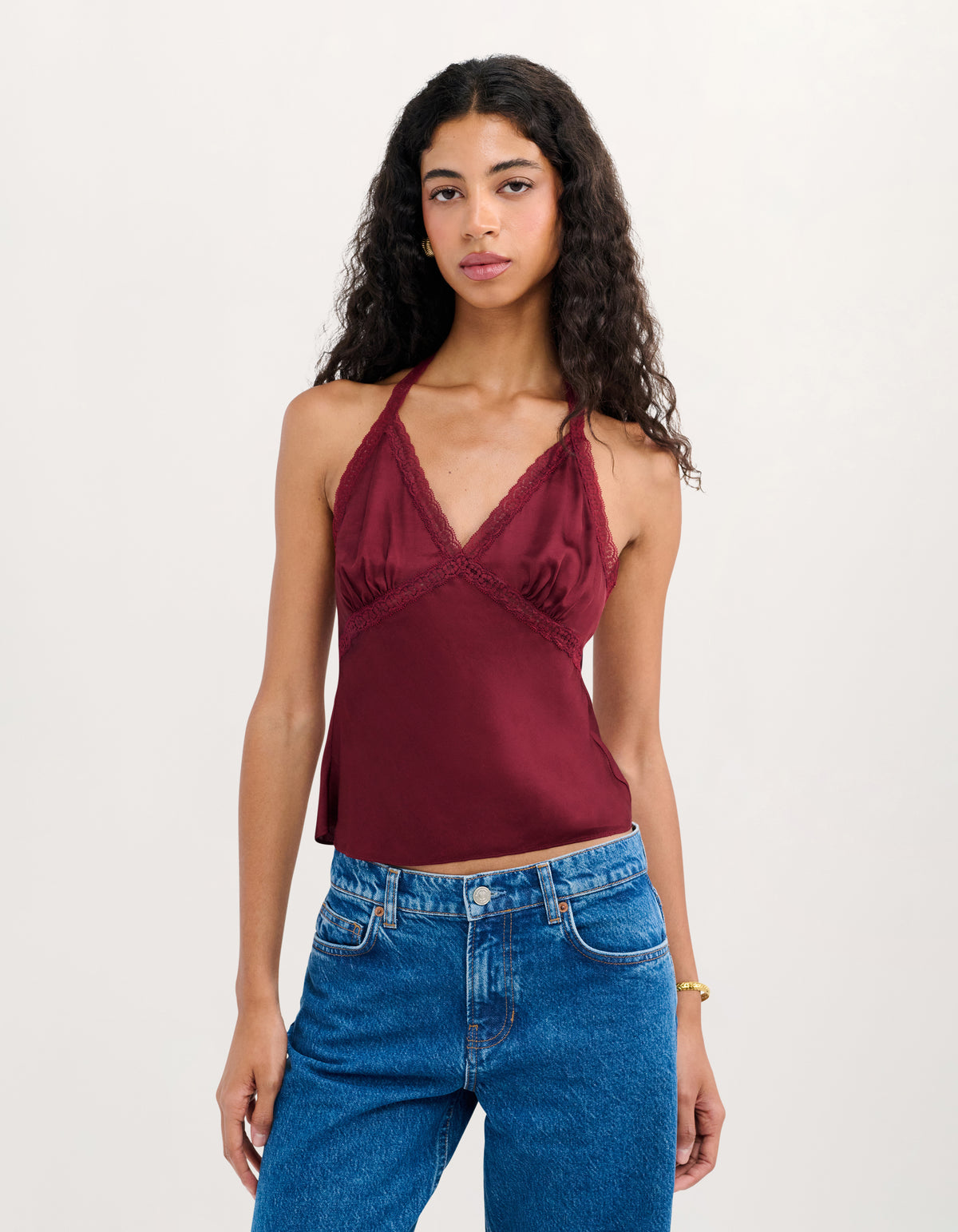 A woman modeling a burgundy satin halter top with lace trim and blue jeans against a plain white studio background.