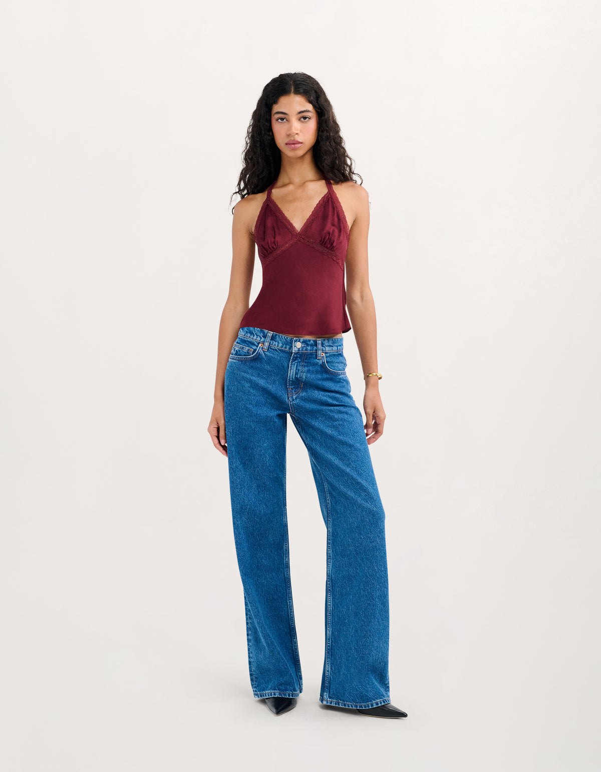 A model stands against a plain light gray studio background wearing a sleeveless burgundy top with ruffle details and high-waisted, wide-leg blue jeans.