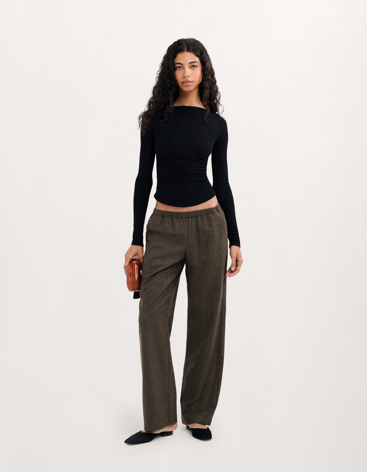 A woman stands against a plain light background wearing a fitted black long-sleeve top and loose dark brown wide-leg pants, with black flats and a small brown clutch.