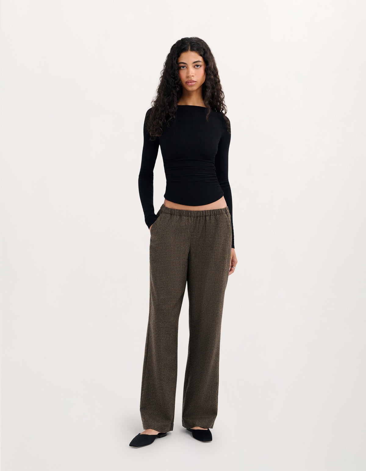 A woman modeling a black long-sleeve ribbed crop top and dark olive brown wide-leg trousers, standing against a plain light gray studio background.